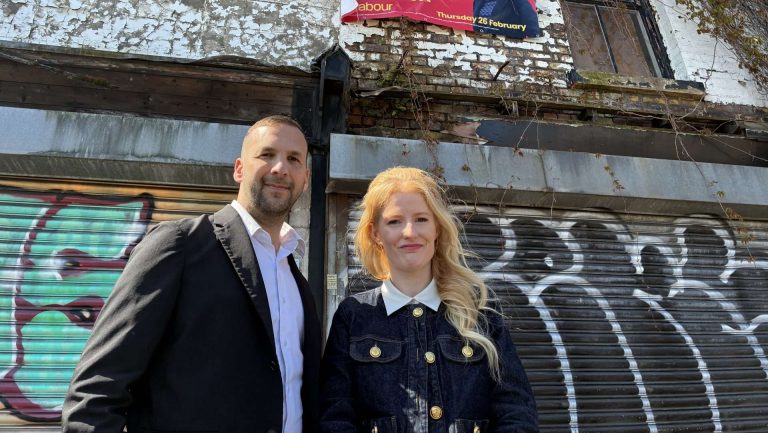Two people in smart clothing stand in front of a graffiti-covered, weathered storefront.