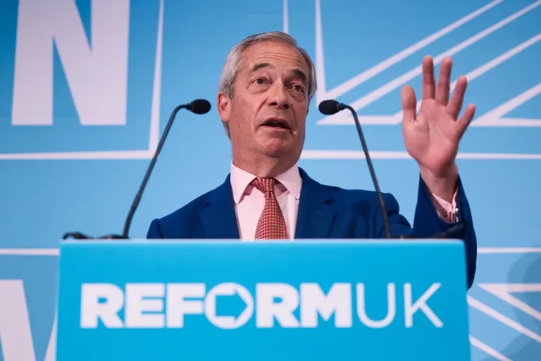 Man in a blue suit and red tie speaks at a Reform UK podium, gesturing with one raised hand behind a blue backdrop.