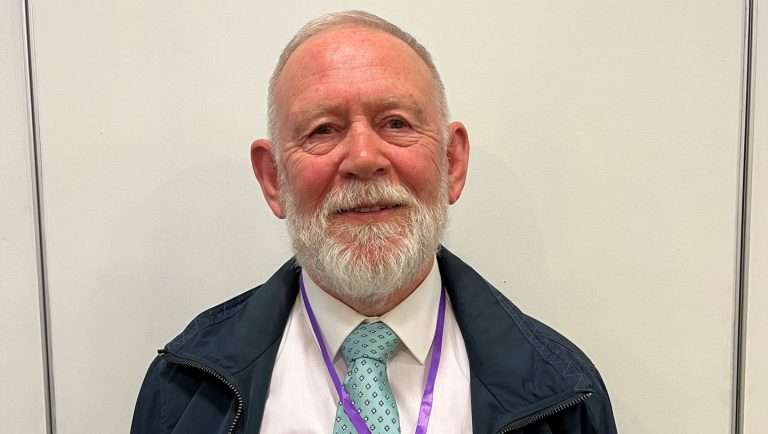 Older man with gray beard in a pink shirt, turquoise patterned tie, navy jacket, and a purple lanyard against a plain wall background.
