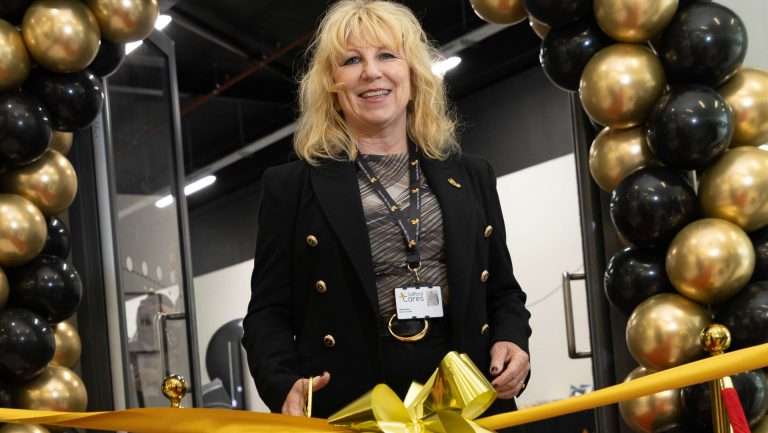 Woman in a black blazer cutting a large yellow ribbon at a balloon arch, smiling during a ceremony.