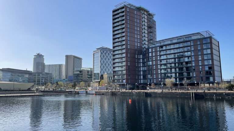 Modern waterfront skyline with tall brick and glass apartment buildings along a calm river.