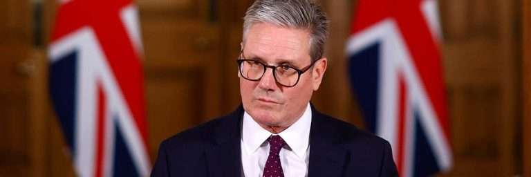 Man in a dark suit and glasses at a formal briefing, with Union Jack flags in the background.