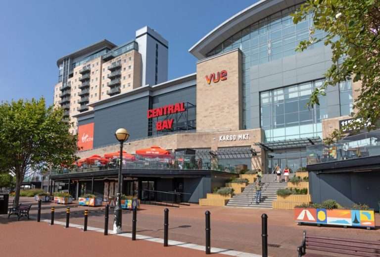 Exterior view of a modern shopping center with 'CENTRAL BAY' and 'vue' signage, outdoor seating under orange umbrellas, on a sunny day
