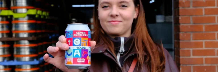 Person holding a colorful can of Seven Brothers beer toward the camera, with brick wall in background.
