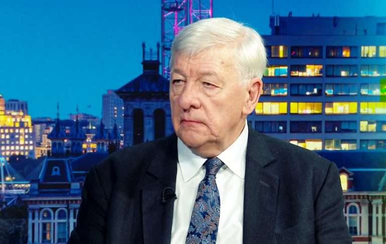 Older man in a suit during an interview against a city skyline backdrop, with a lapel mic visible on his jacket.