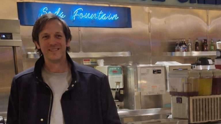 Smiling man in a navy jacket poses at a stainless steel counter inside an ice cream shop with festive wreath on a grill nearby.