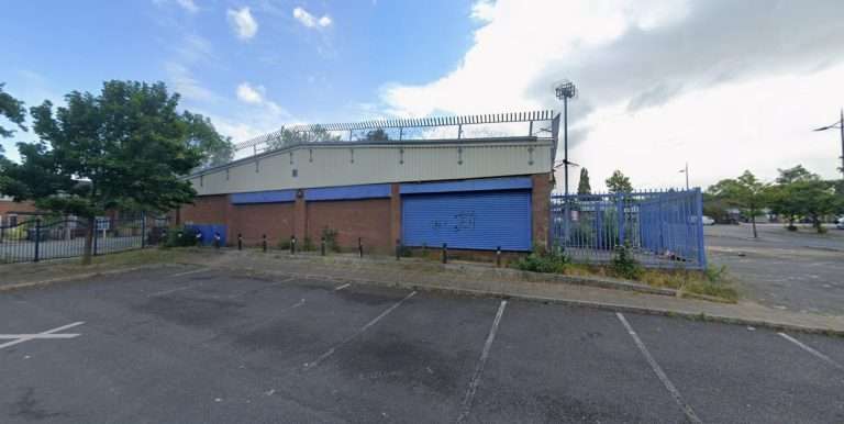 Industrial brick building with blue roll-up doors and a barbed-wire fence on top, in a paved parking area.