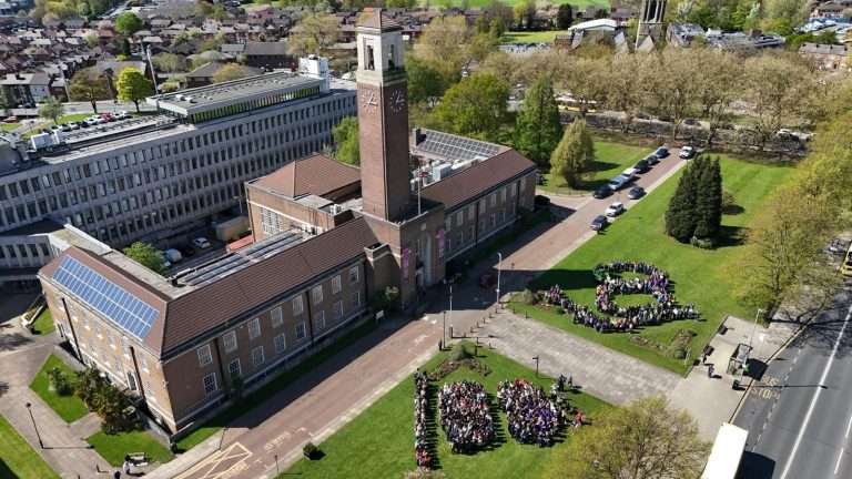 Aerial view of a brick university building with a tall clock tower, surrounded by a lawn where people form circular groups on the grass.