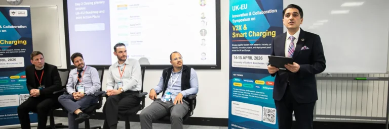 Panel discussion at a conference: four men seated as speakers, one man in a suit presenting with a tablet, banners reading 'UK-EU Innovation & Collaboration' and 'V2X & Smart Charging' in a classroom stage setting.