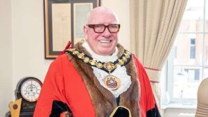 Older man in ceremonial red robe with fur trim and gold chain, smiling and holding a black tricorn hat in a formal office.