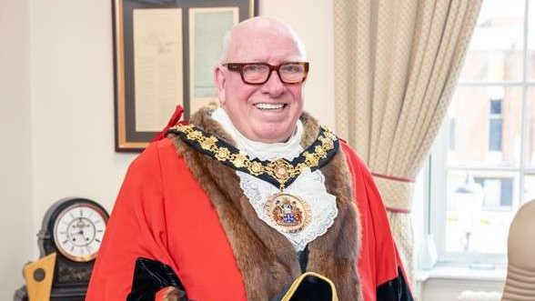 Older man in ceremonial red robe with fur trim and gold chain, smiling and holding a black tricorn hat in a formal office.