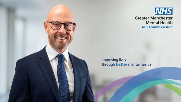 Smiling man in a navy suit and glasses standing next to an NHS Greater Manchester Mental Health banner.