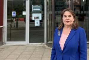 Woman in a bright blue blazer and pink top standing outside glass doors, facing the camera in a public building entrance.