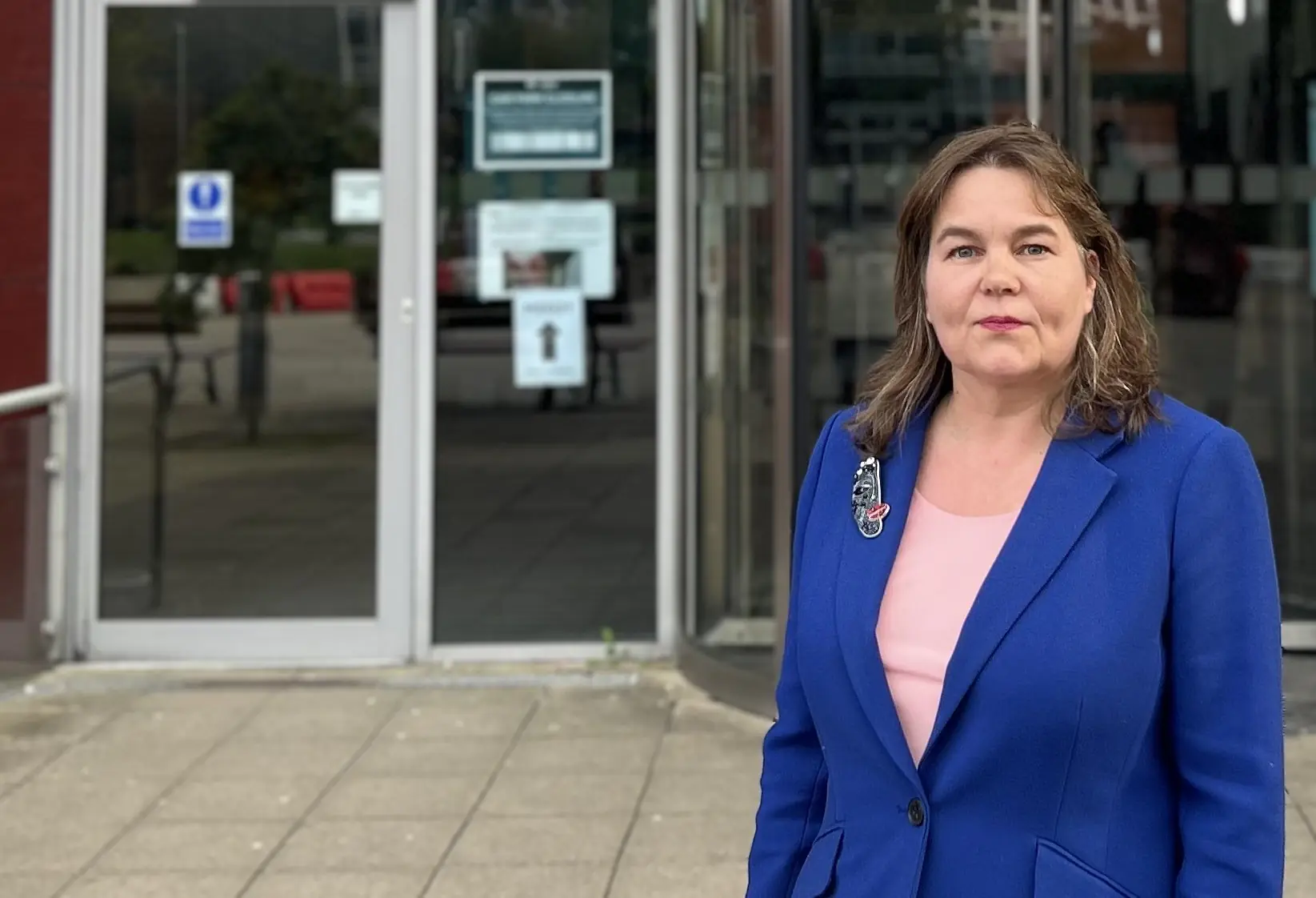 Woman in a bright blue blazer and pink top standing outside glass doors, facing the camera in a public building entrance.