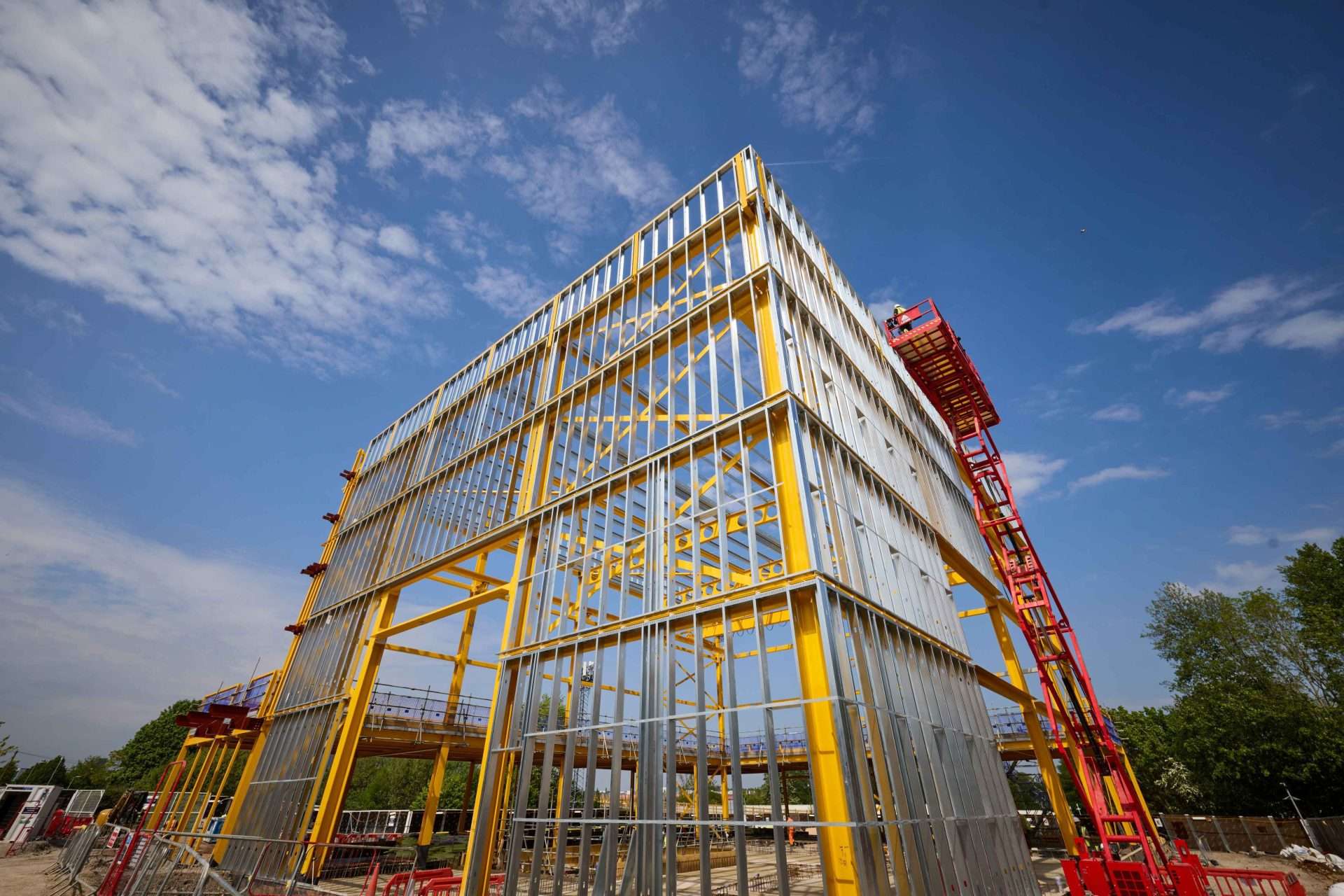 Yellow steel-framed building under construction against a blue sky with a red lifting platform nearby