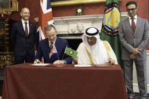 Two men sign documents at a formal ceremony, flanked by flags and two observers in suits in an ornate room.