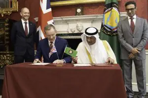 Two men sign documents at a formal ceremony, flanked by flags and two observers in suits in an ornate room.