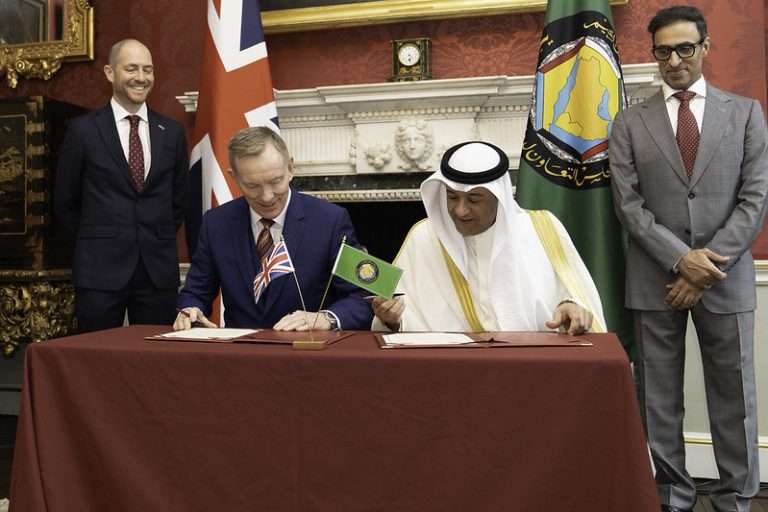 Two men sign documents at a formal ceremony, flanked by flags and two observers in suits in an ornate room.