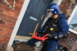 Police officer in blue gear with helmet and visor at a brick front door, holding a red tool amid debris outside the entryway.