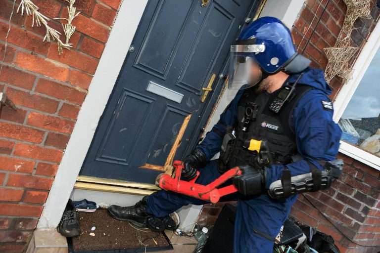 Police officer in blue gear with helmet and visor at a brick front door, holding a red tool amid debris outside the entryway.