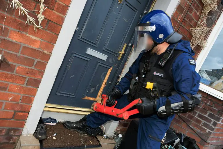 Police officer in blue gear with helmet and visor at a brick front door, holding a red tool amid debris outside the entryway.
