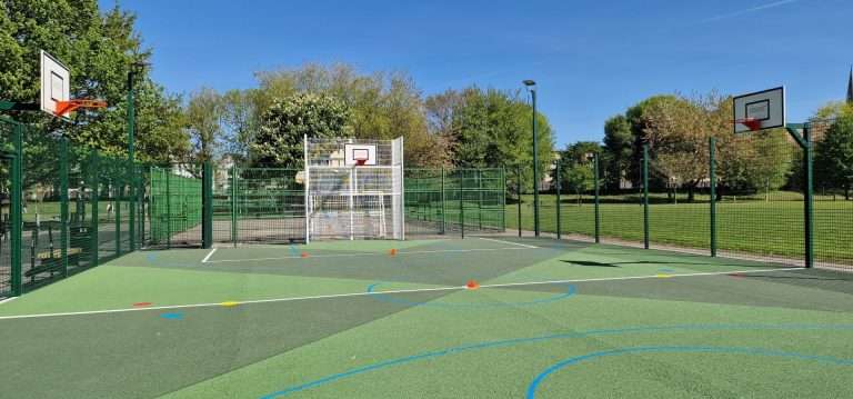 Outdoor fenced basketball court in a park, two hoops, green surface with blue and white lines, and orange cones setup on the court under a clear blue sky.