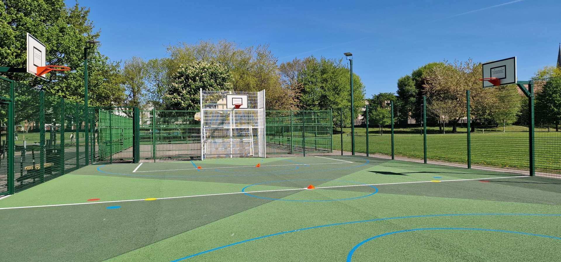 Outdoor fenced basketball court in a park, two hoops, green surface with blue and white lines, and orange cones setup on the court under a clear blue sky.