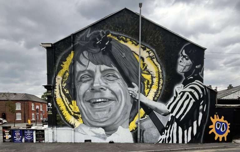 Giant mural on a building showing a grayscale portrait of a smiling woman with a yellow halo, and a black-and-white striped figure to the right; an orange sun icon appears bottom-right.