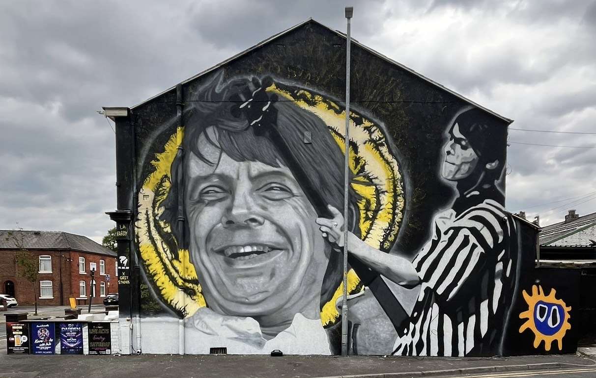 Giant mural on a building showing a grayscale portrait of a smiling woman with a yellow halo, and a black-and-white striped figure to the right; an orange sun icon appears bottom-right.