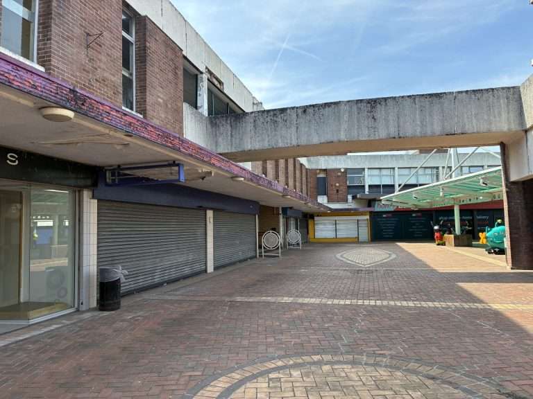 Empty outdoor shopping arcade with closed storefronts, brick buildings, and a concrete overhead walkway under a blue sky.