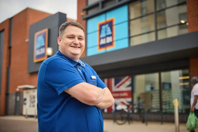 Smiling man in a blue polo with arms crossed outside a brick-and-glass storefront above him.