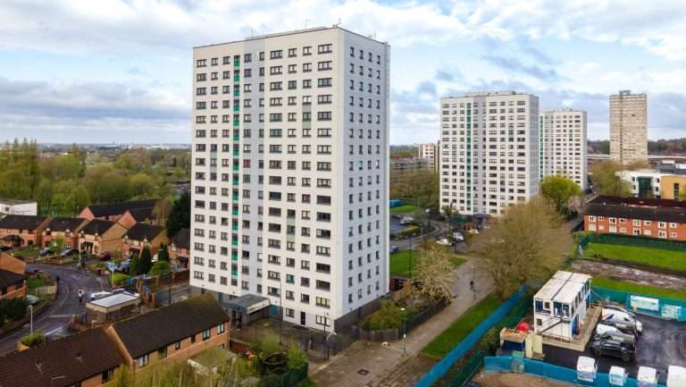High-angle view of several tall white apartment blocks in a suburban area with trees and roads under a blue cloudy sky.