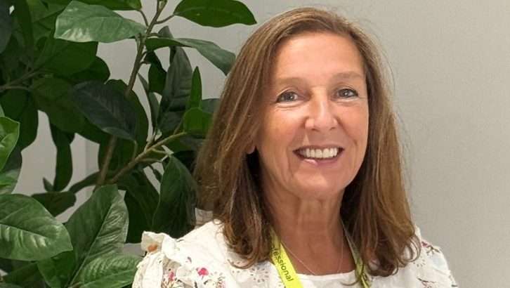 Smiling woman with light brown hair wearing a floral blouse and a yellow lanyard, indoors beside a green plant.