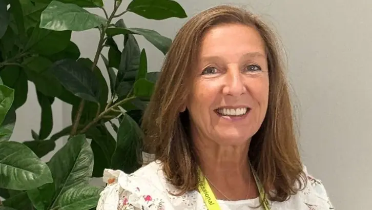 Smiling woman with light brown hair wearing a floral blouse and a yellow lanyard, indoors beside a green plant.
