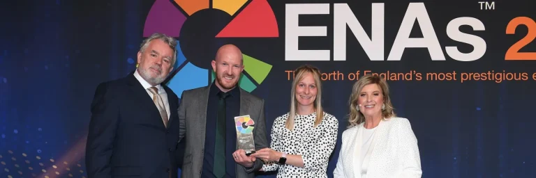 Four adults in business attire pose on a stage for a photo at an ENAS awards event; the man second from the left holds a glass trophy with a multicolor circular emblem, with the ENAS logo behind them.