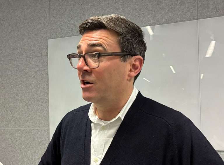 Side profile of a man wearing brown glasses, a white shirt, and a dark cardigan in an office setting behind glass panels.
