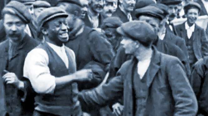 Two men shake hands in a crowded rail of workers wearing caps and vests, suggesting a scene of camaraderie or solidarity.