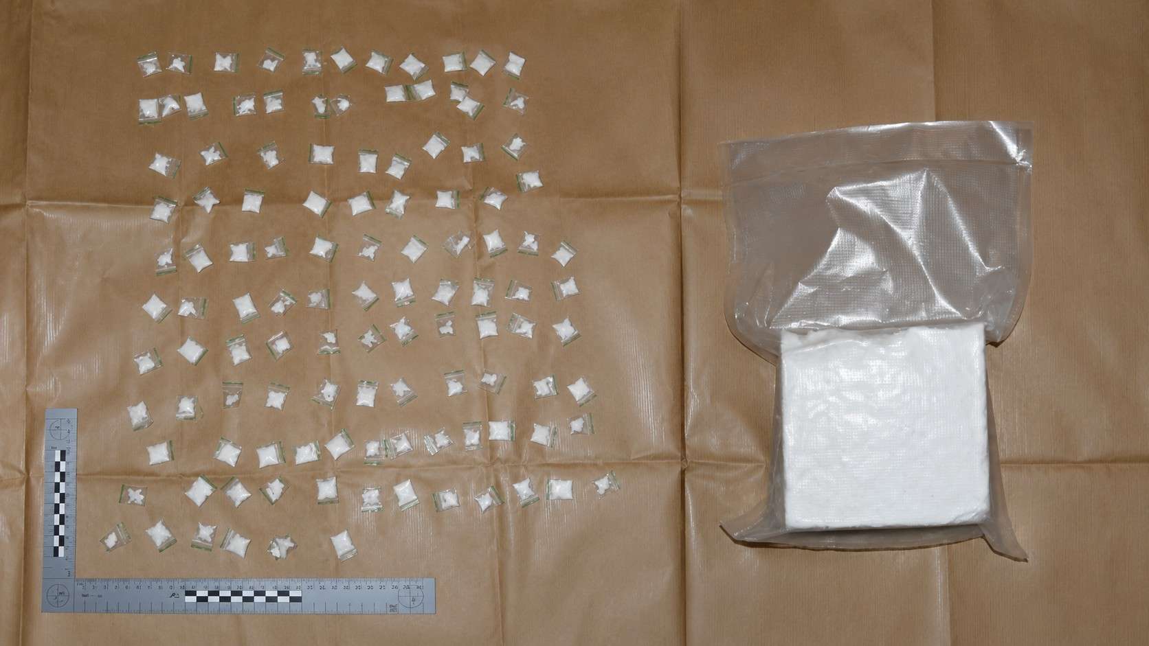 Assorted small sealed plastic bags containing a white crystalline substance scattered on brown paper, with a gray vacuum-sealed bag and a white pad nearby for scale and evidence.