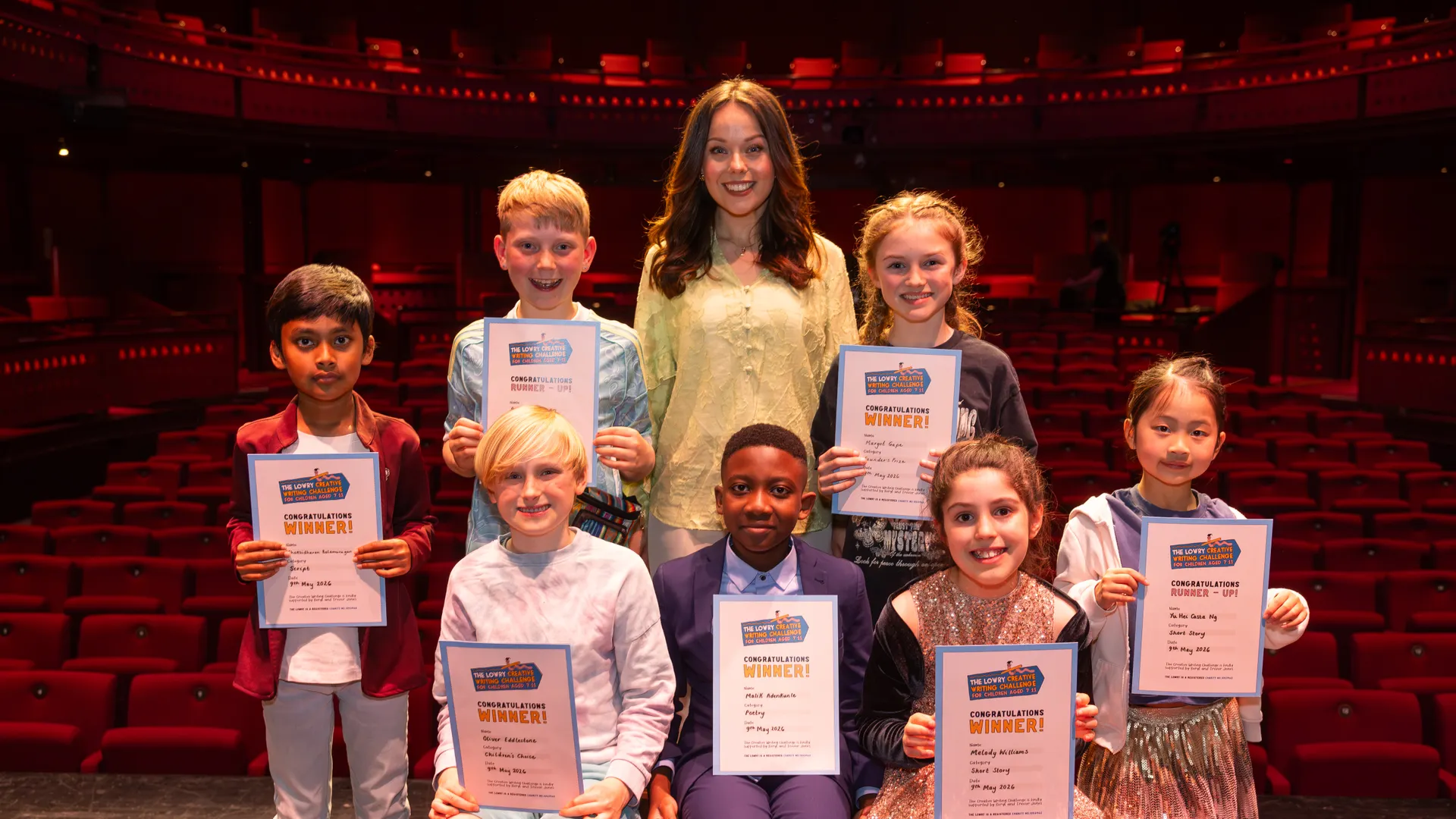 Group of smiling children holding certificates on a theater stage with red seats in the background, led by a woman instructor/host.