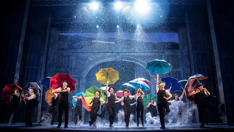Ensemble of performers on a stage, each holding colorful umbrellas as artificial rain falls under bright blue spotlights.