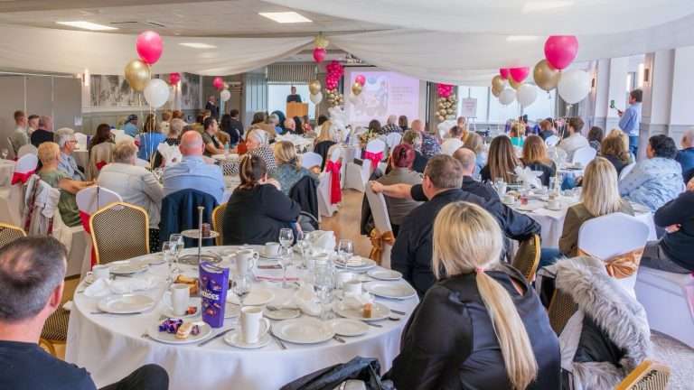 People seated at round tables in a banquet hall, listening to a speaker on a stage with pink and gold balloons overhead