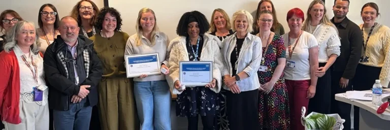 Group of diverse people posing together in a room; two women in front hold certificates, smiling at the camera.