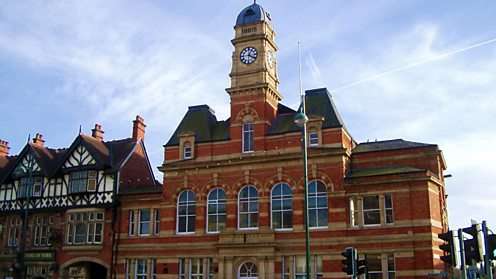 Brick town hall with a clock tower and arched windows, flanked by Tudor-style buildings under a blue sky.