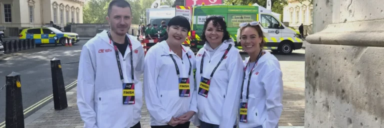 Four volunteers in white jackets with finish lanyards pose on a city street near the finish line, with ambulances and support crews in the background.