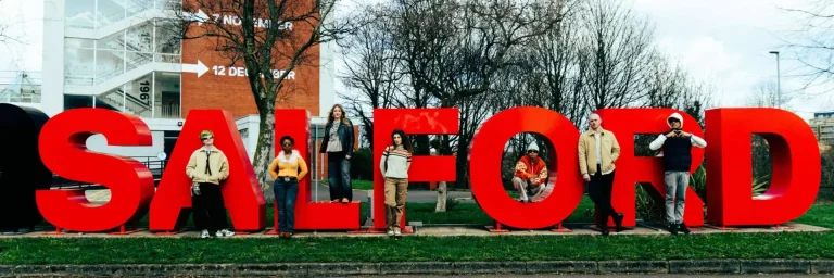 Group of diverse people posing beside oversized red letters outdoors on a campus-like lawn.