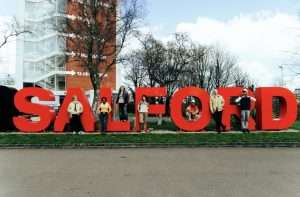 Group of people posing with oversized red letters spelling 'SALFORD' outdoors in a grassy area, with trees and a modern building behind.