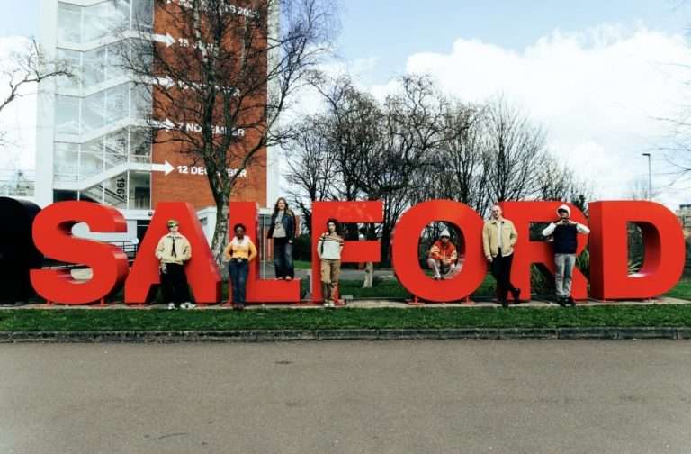 Group of people posing with oversized red letters spelling 'SALFORD' outdoors in a grassy area, with trees and a modern building behind.