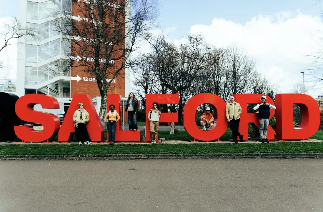 Group of people posing with oversized red letters spelling 'SALFORD' outdoors in a grassy area, with trees and a modern building behind.