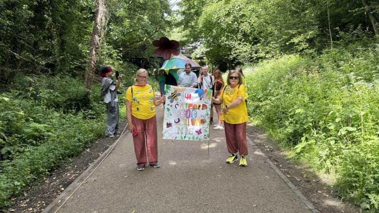 Group of people in a park parade holding a colorful banner, with a rainbow tent behind them.