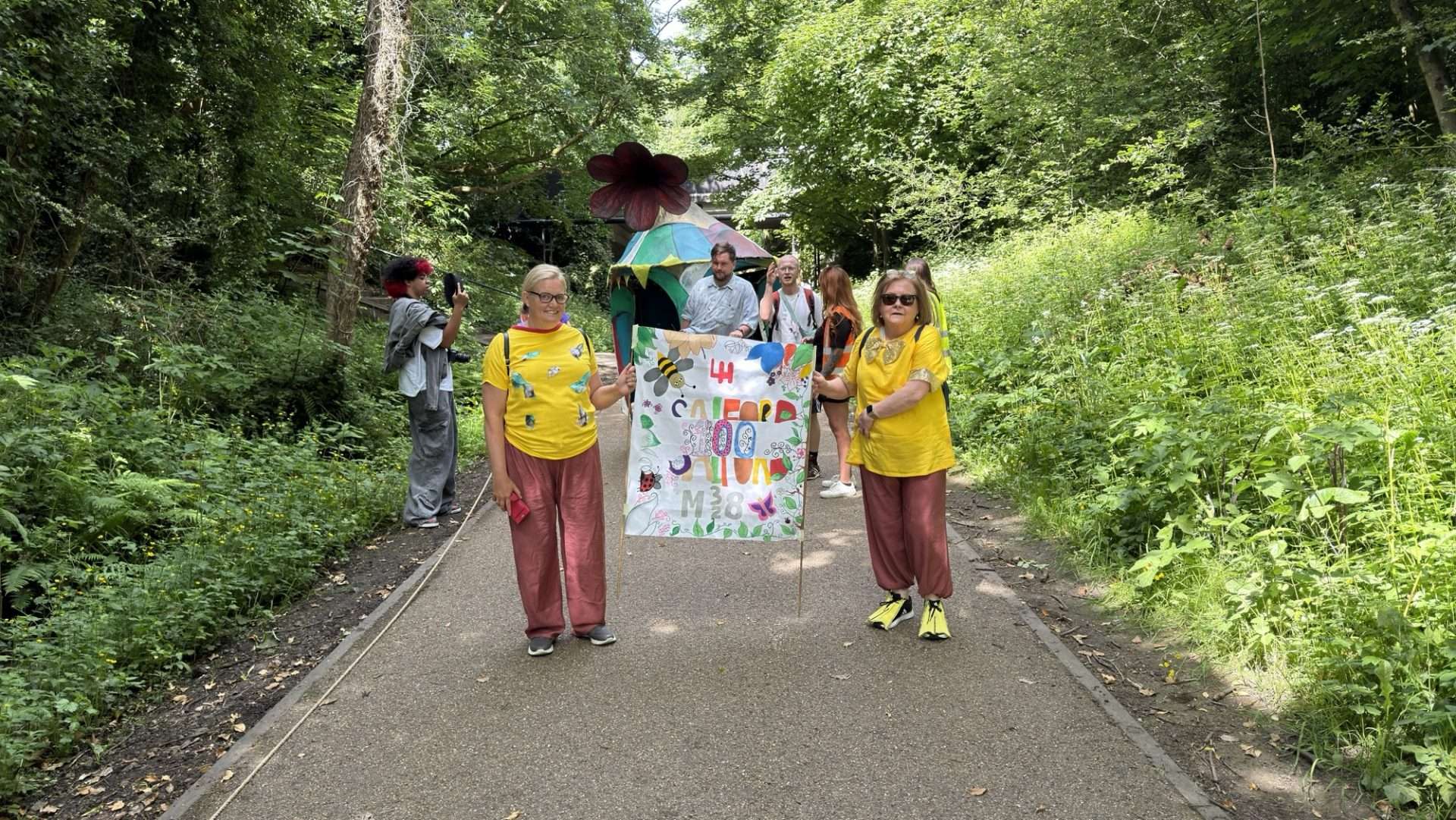 Group of people in a park parade holding a colorful banner, with a rainbow tent behind them.